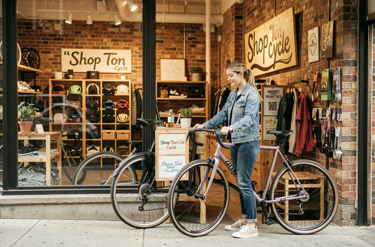 Jeune femme examinant un vélo urbain fixie dans la boutique indépendante Shop Ton Cycle, décor en briques apparentes et esprit artisanal